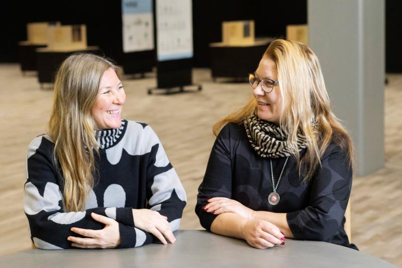 HR-partneri Paula Jalo ja varhaiskasvatusjohtaja Miia Kemppi nojaavat kyynerpäillä pöytään vierekkäin ja katsovat toisiinsa nauraen.
