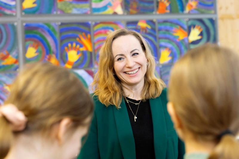 Rehtori Mari Koivukangas seisoo värikkään seinän edessä ja hymyilee iloisesti. Etualalla on kaksi oppilasta selkä kuvaan päin.