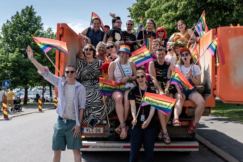 Joukko iloisia ihmisiä istuu rekan lavalla. He heiluttavat Helsinki Pride -viirejä.