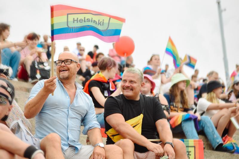 Ihmisiä istumassa kirkon portailla Pride-lippujen kanssa. Etualalla on kaksi miestä, jotka nauravat. Toinen pitää Helsinki-logolla varustettua sateenkaarilippua kädessä ja katsoo vasemmalle.
