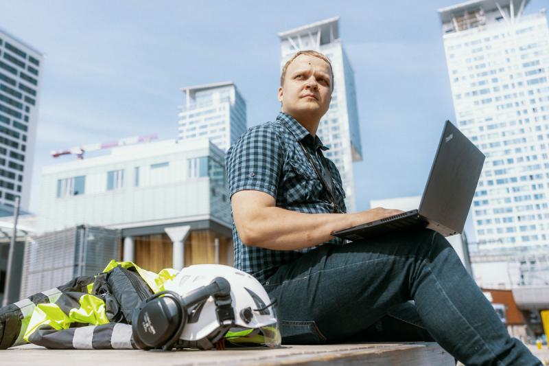 Mies istuu ulkona kannettava tietokone sylissään ja rakennusmiehen takki ja kypärä vieressään. Takana näkyvät Kalasataman korkeat kerrostalot.