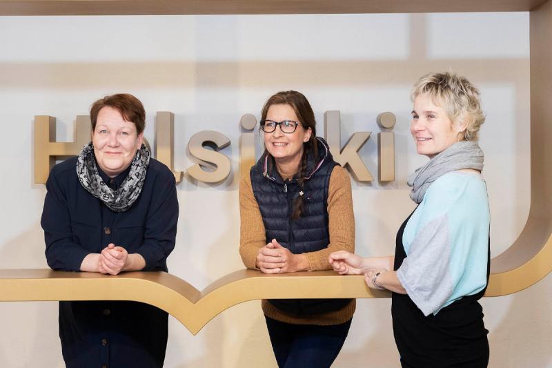 Johtava ohjaaja Soile Härkönen, pehtoori Tiina Raitoja ja päiväkodinjohtaja Inka Railo nojaavat kyynerpäillä suuren Helsinki-logon reunukseen ja hymyilevät.
