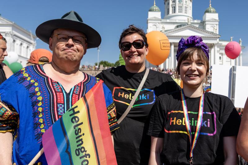 Pormestari Juhana Vartiainen ja kaksi naista ovat pukeutuneet värikkäästi. Naisilla on Helsinki Pride -paidat ja kaulanauhat. Juhanan kädessä on Helsinki Pride -viiri. He seisovat Senaatintorilla ja hymyilevät.